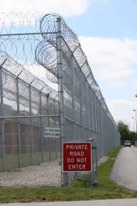 2 layers of high security fence with Concertina wire strung on top. Two signs are attached to the fence, one small white one that reads "No pedestrian traffic allowed" and a large red one that reads "private road do not enter"