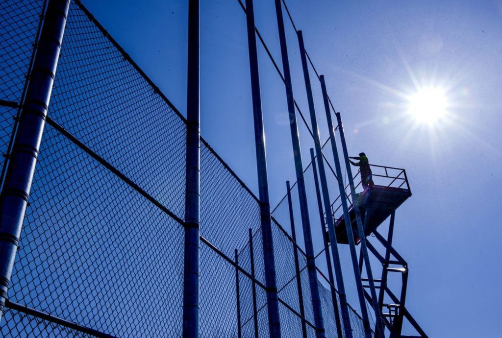 An American Fence Company worker installing chain link fabric on Nebraska Wesleyan University's backstop. He is standing on a lift and has the sun blazing behind him in a cloudless blue sky