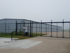 A black vinyl chain link cantilever gate with barb wire installed in front of a large nondescript warehouse. This hate has a manual keypad installed that is wired to an access control operator