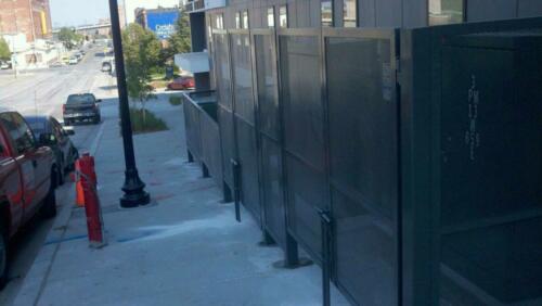6 - 8 foot black woven and welded wire fence creating an enclosure around outdoor power boxes on cement along sidewalk