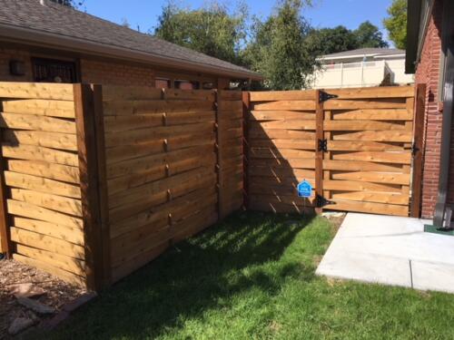 5-8 foot tall horizontal wooden basket weave fence with vertical slightly off-colored fence posts and similarly styled single manual swing gate