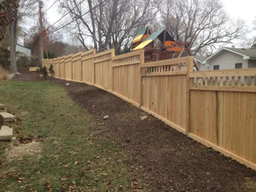 5-8 foot tall decorative wooden privacy fence staggering height (no ground to baseline gap) due to hilly grassy terrain with vertical about 2-5 inch tall show/hide gaps occurring along the top of fence with an additional horizontal plank they above them. 