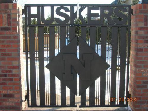 6-8 foot flat rectangular black iron ornamental double swing gate connecting to brick columns with the word "HUSKERS" along the top and a large emblem in the center of gate with husker logo