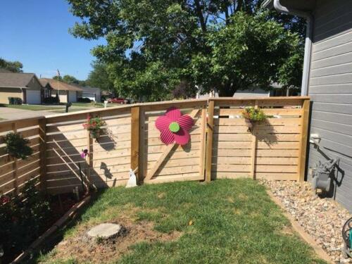 6-8 foot tall horizontal wooden fence with off-colored post with inward overhang and plants hanging on fence gaps