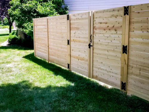 6-8 foot tall wooden horizontal planks with vertical off-colored fence posts with similar single manual swing gate