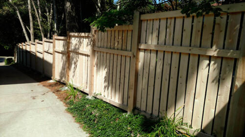 6-8 tall vertical semi-private wooden fence with staggered top alignment due to uneven terrain, also has forward horizontal plank 1 - 1 1/2 feet beneath the top of fence line. Baseline is 1-4 inches above ground level due to staggered height