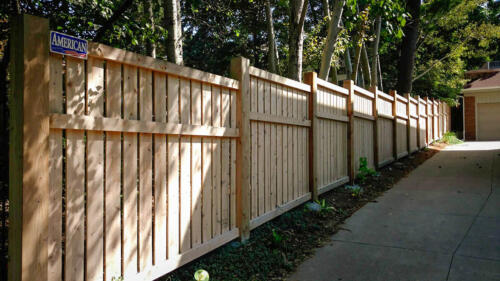 6-8 tall vertical semi-private wooden fence with staggered top alignment due to uneven terrain, also has forward horizontal plank 1 - 1 1/2 feet beneath the top of fence line. Baseline is 1-4 inches above ground level due to staggered height