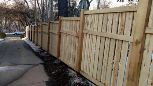 6-8 tall vertical semi-private wooden fence with staggered top alignment due to uneven terrain, also has forward horizontal plank 1 - 1 1/2 feet beneath the top of fence line. Baseline is 1-4 inches above ground level due to staggered height