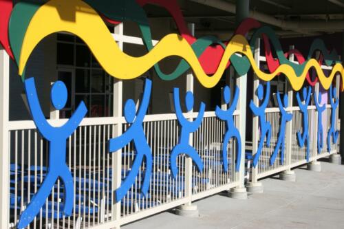 Railing with large decorative blue stick figures playing and wavy banner (red, green, yellow) above figures