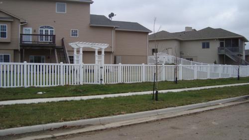 4-6 foot tall white fencing with a 4-8 inch gap between boards. There is a boxed white archway with a gate underneath, located towards the center of the fence.