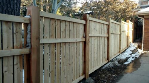 6-8 tall vertical semi-private wooden fence with staggered top alignment due to uneven terrain, also has forward horizontal plank 1 - 1 1/2 feet beneath the top of fence line. Baseline is 1-4 inches above ground level due to staggered height